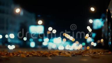 城市灯光和汽车在交通背景下行驶。 秋天的树叶从路过的汽车上随风飘散在夜路上。